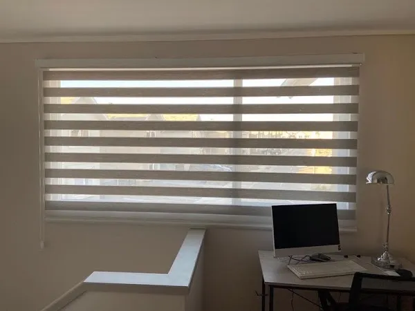 Roller duo con franjas alternadas regulando la luz sobre un escritorio junto a la ventana