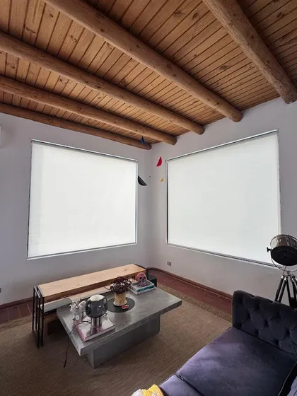 Living con cielo de vigas de madera y dos rollers claros bajados sobre los ventanales